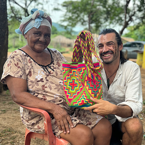 Fotografía de una mujer sentada y sonriendo hacia la cámara, junto a Christian Byfield quien sostiene un bolso wayúu que compró en su tour de 3 días en La Guajira