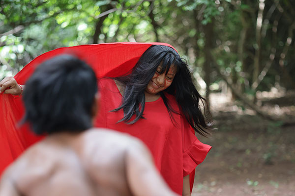 Mujer wayúu tiene puesto un vestido rojo frente a un joven wayúu que está de espaldas a la cámara sin camisa, los cuales se encuentran en uno de los resguardos que hacen parte del tour por el Cabo de la Vela y Punta Gallinas