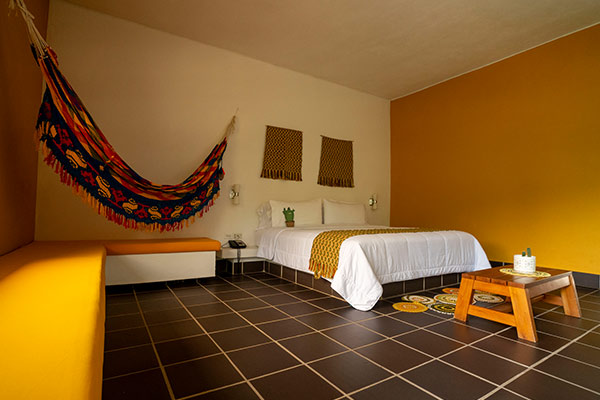 Habitación de hotel con una cama doble, una hamaca, una mesita de madera, y sillas esquineras de tonalidades blancas y amarillas, que hace parte de los paquetes en oferta para ir a La Guajira